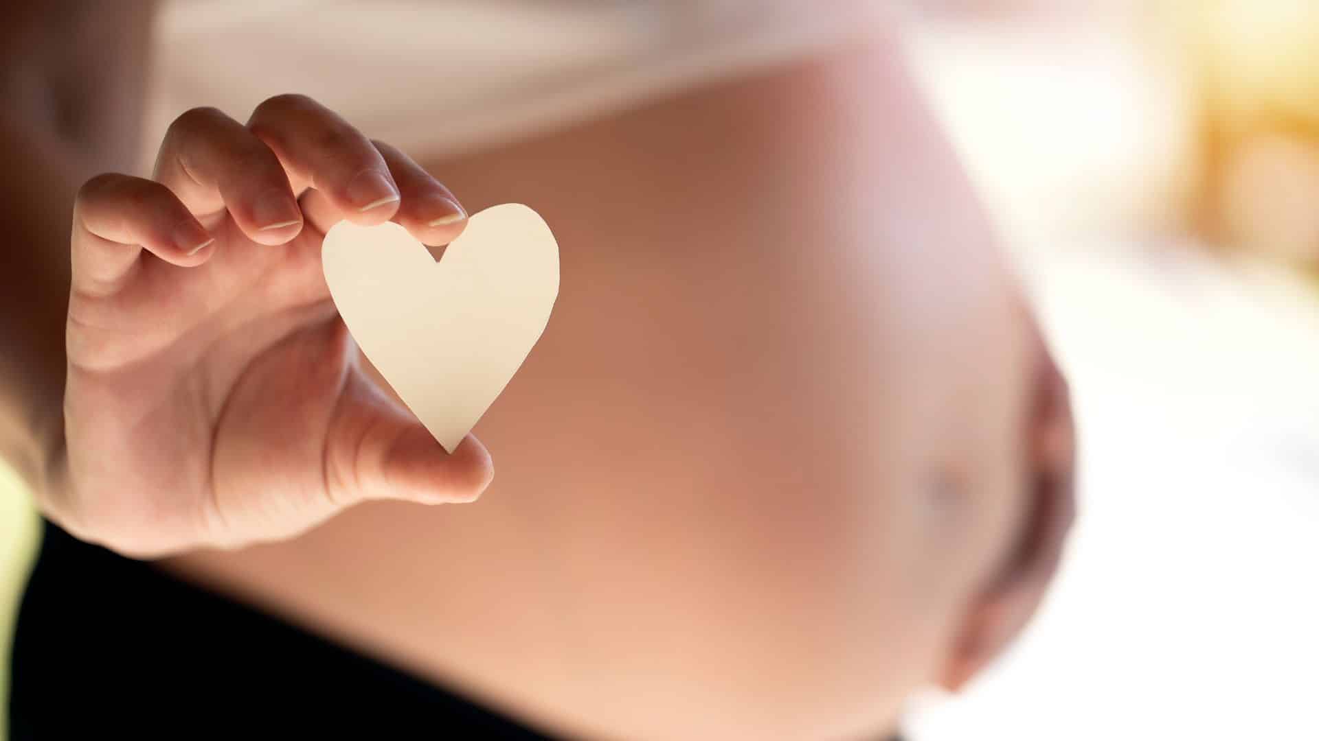 Ventre de femme enceinte avec un coeur blanc en papier dans la main