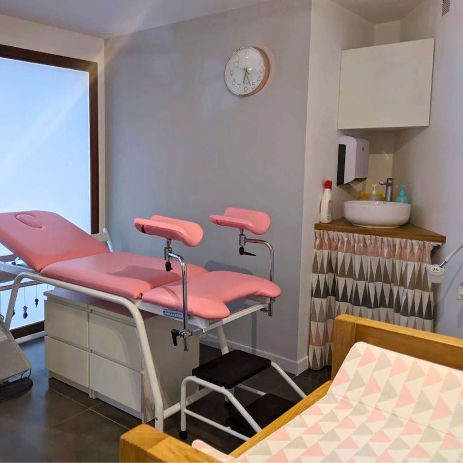 Table d’examen gynécologique rose dans un cabinet médical propre avec lavabo et équipements d’hygiène, ambiance rassurante et soignée.